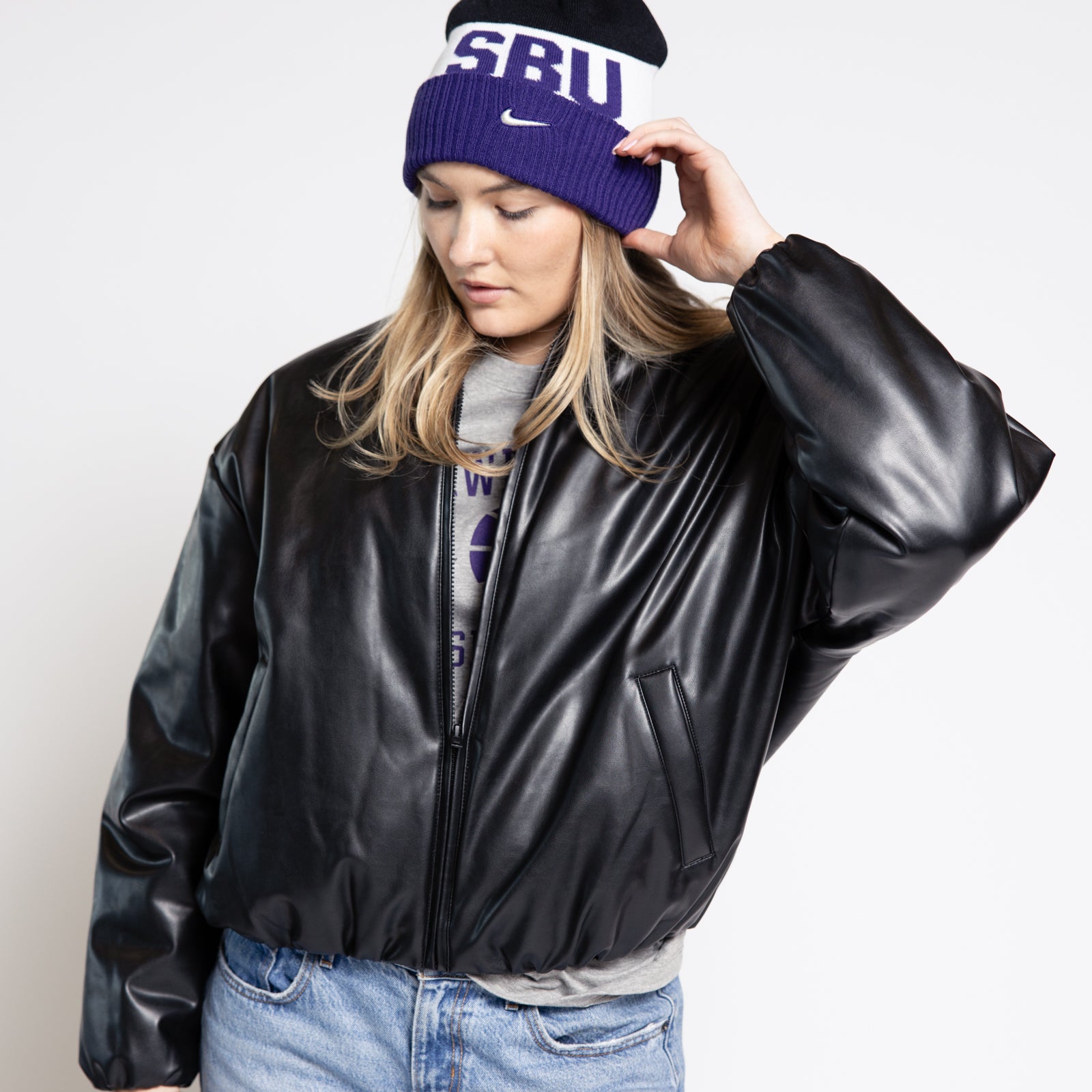 Woman wearing a black leather jacket, blue jeans, and a purple beanie with 'SBU' text on a white background.