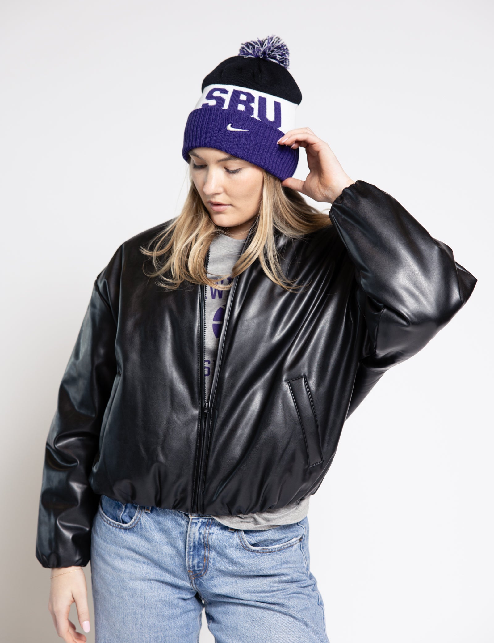 Woman wearing a black leather jacket, blue jeans, and a purple beanie with 'SBU' text on a white background.