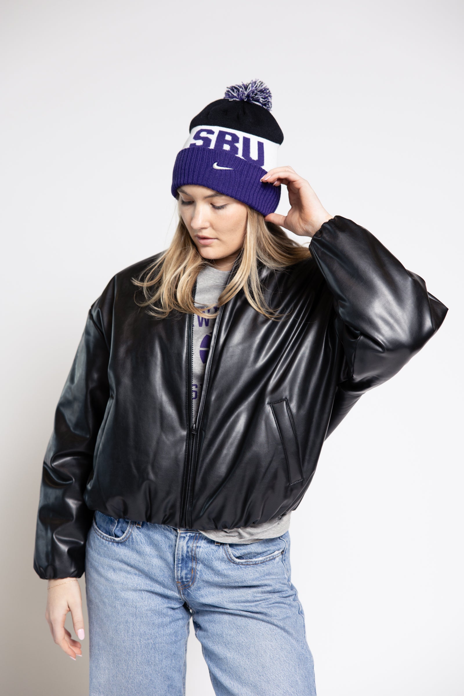 Woman wearing a black leather jacket, blue jeans, and a purple beanie with 'SBU' text on a white background.