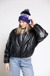 Woman wearing a black leather jacket, blue jeans, and a purple beanie with 'SBU' text on a white background.